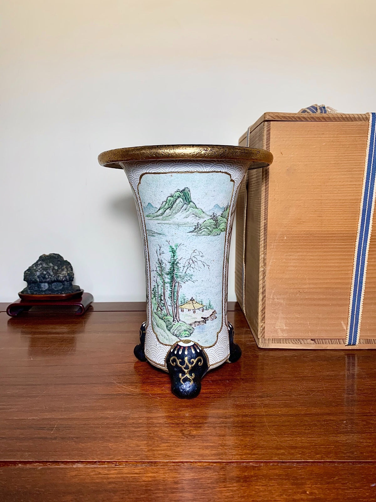 A tall, white glazed pot with a painted mountain scene and a gold rim.