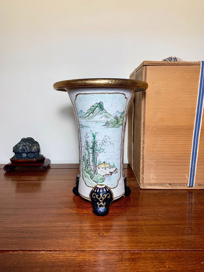 A tall, white glazed pot with a painted mountain scene and a gold rim.