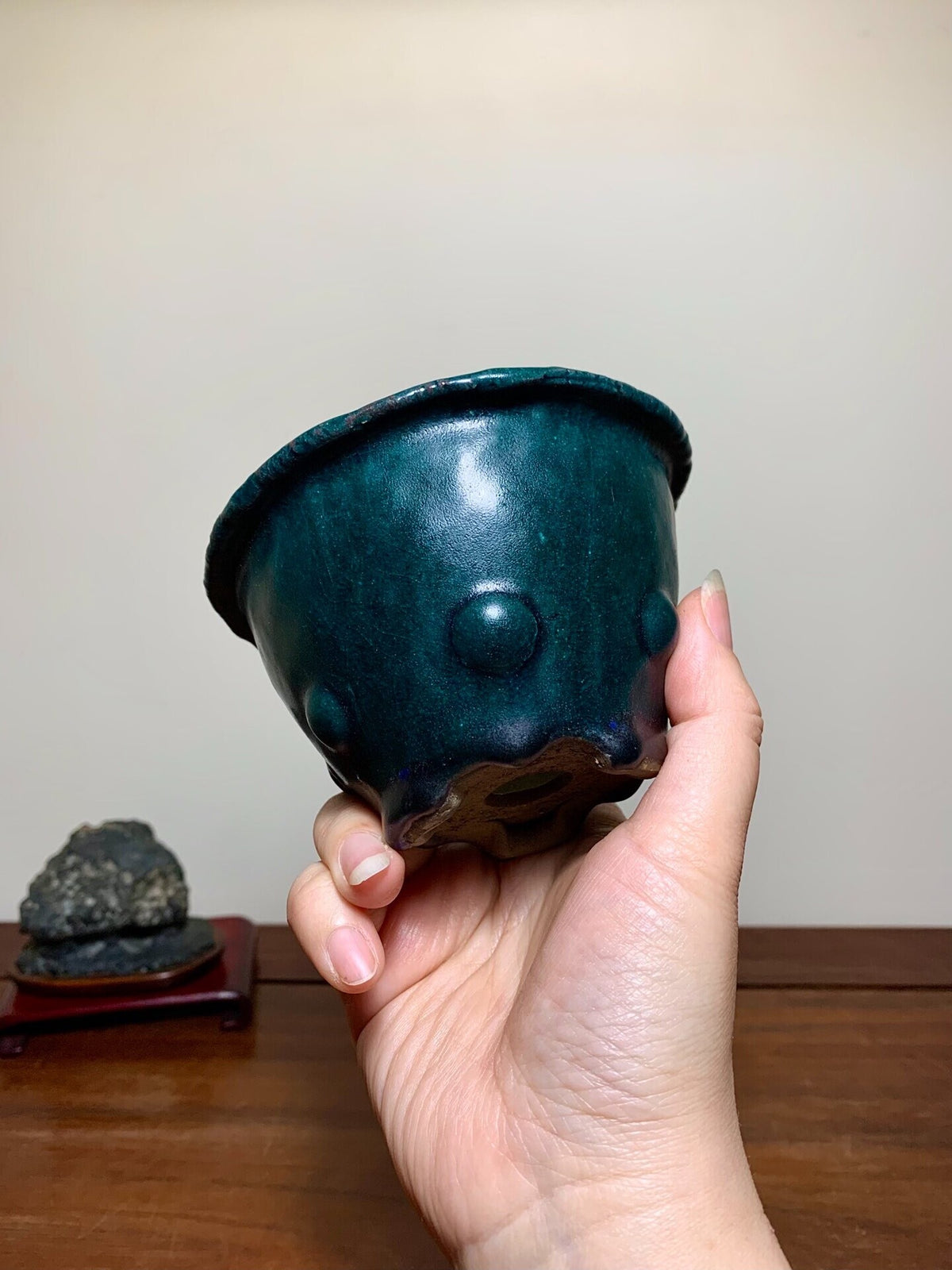 A hand holds a glossy, dark teal ceramic pot with three raised bumps and a scalloped rim.
