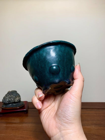 A hand holds a glossy, dark teal ceramic pot with three raised bumps and a scalloped rim.