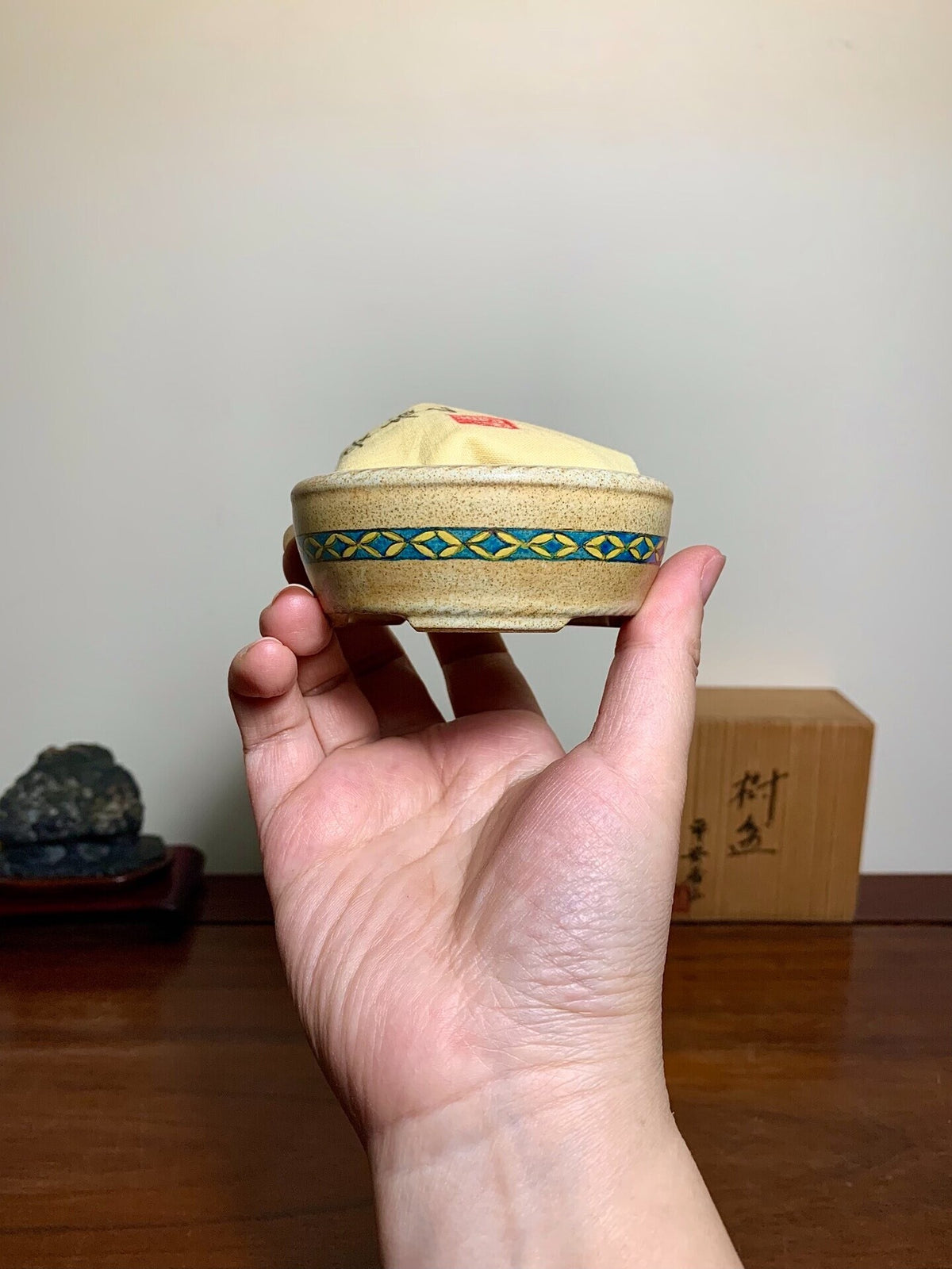 A hand holds a small, beige, unglazed pot with a blue and yellow geometric band.
