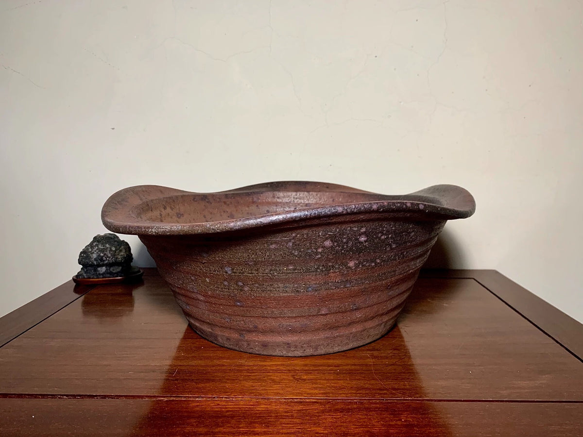 A dark brown, wavy-rimmed ceramic pot with horizontal ridges and speckled glaze sits on a wooden surface.
