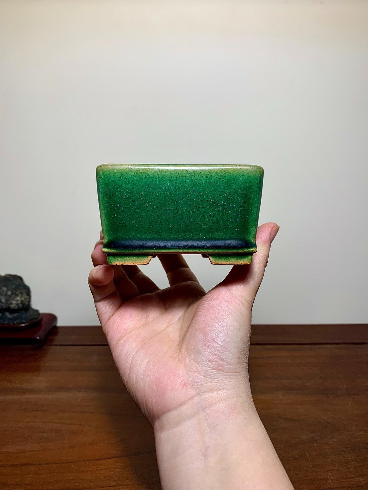 A hand holds a small, square green glazed pot with a darker rim and unglazed base.