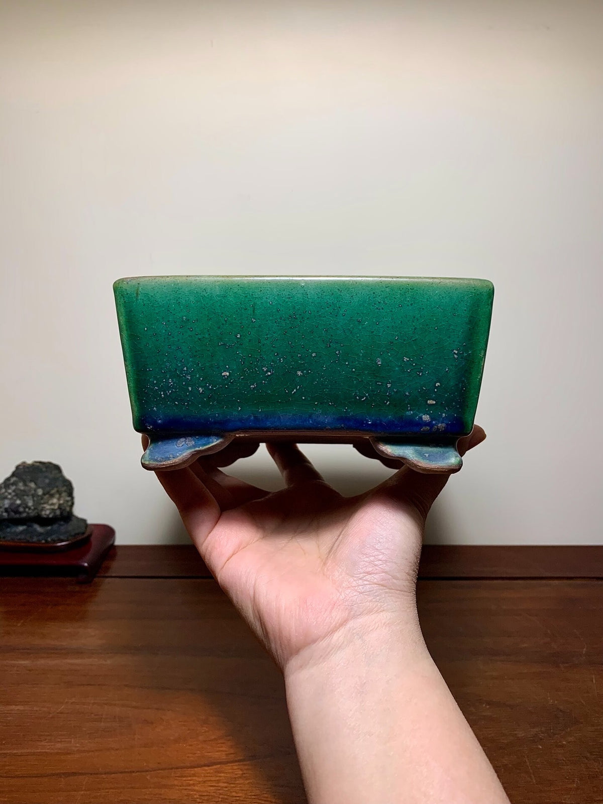 A hand holds a square green glazed pot with a speckled finish and blue rim.