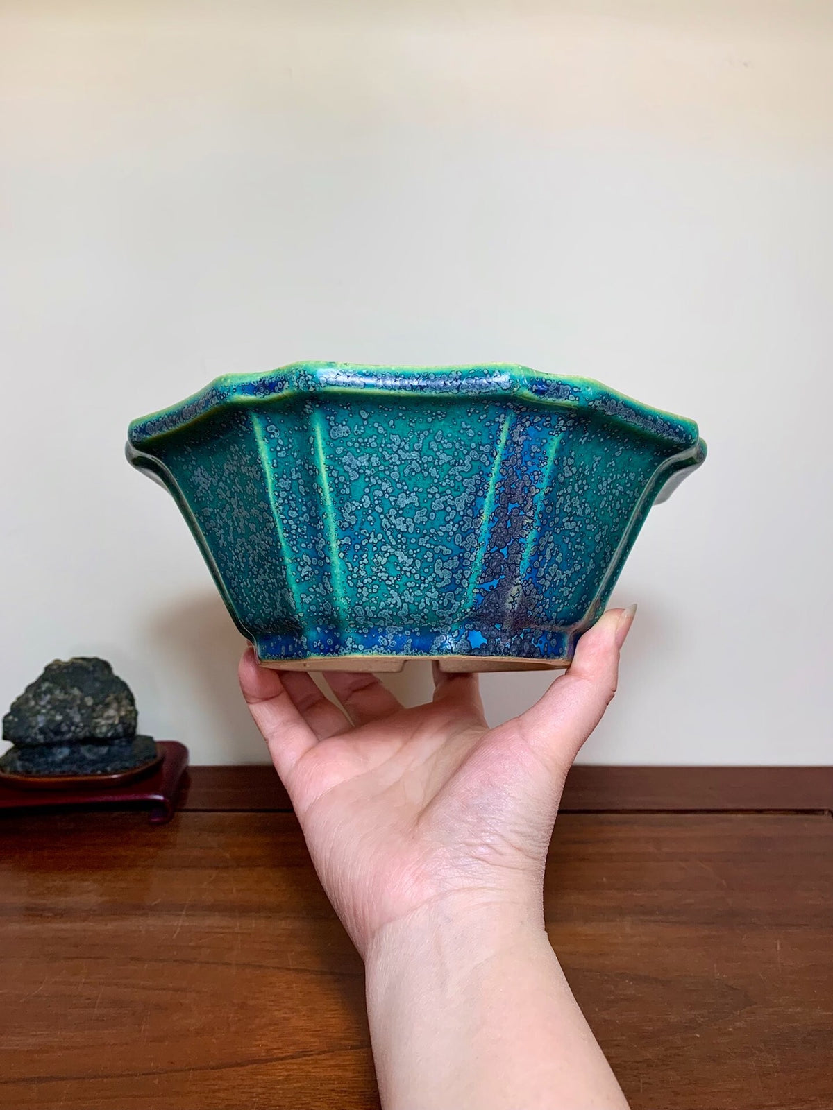 A hand holds a glossy, teal-glazed ceramic pot with a scalloped rim and vertical ridges.