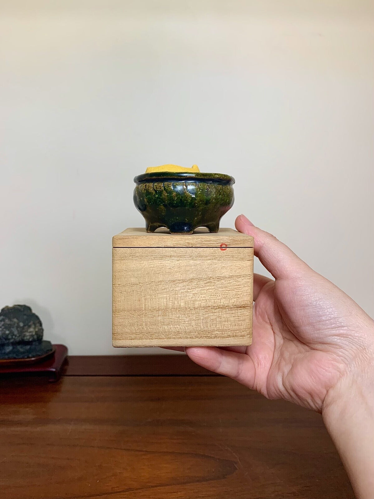 Hand holds small, dark green glazed pot with yellow rim, atop light wood box.
