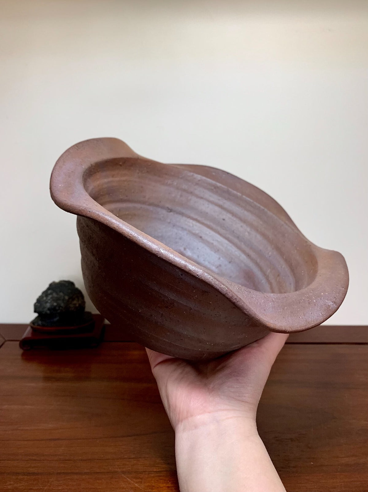 A hand holds a brown, unglazed ceramic pot with a wide rim and visible horizontal ridges.