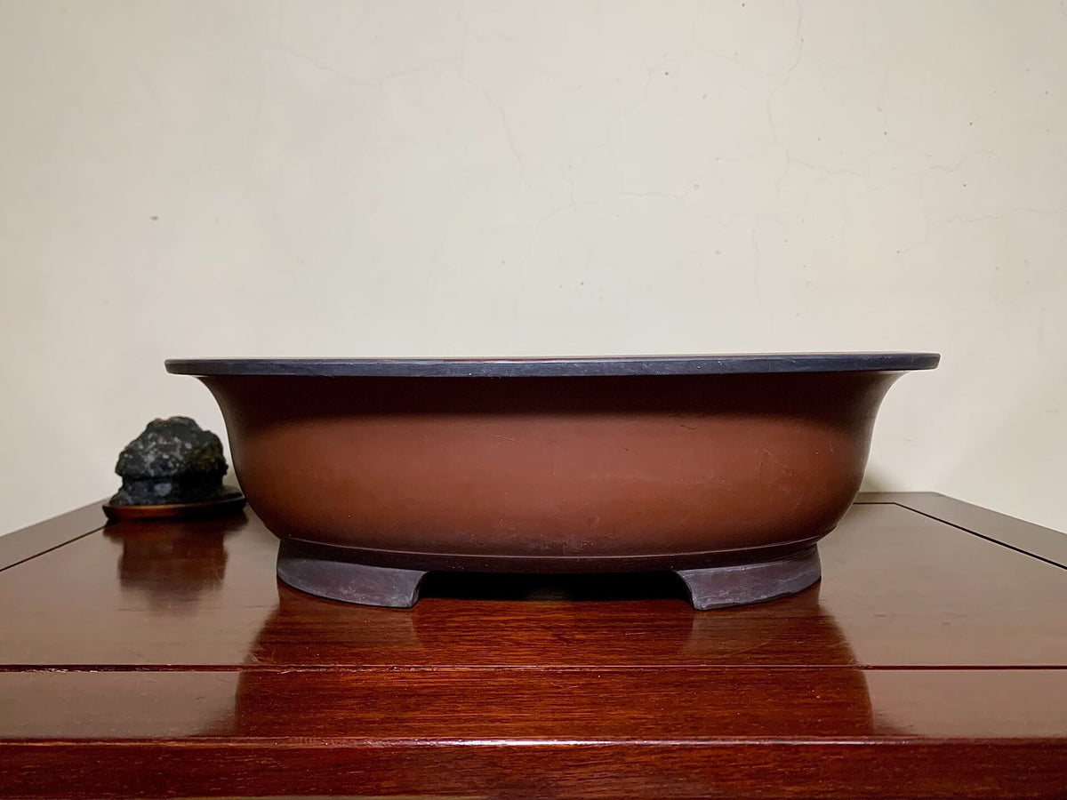 A glossy brown ceramic pot with a dark rim and three feet, beside a small dark rock.