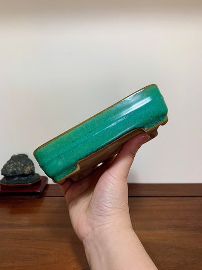 A hand holds a glossy, rectangular green glazed pot with a brown rim.