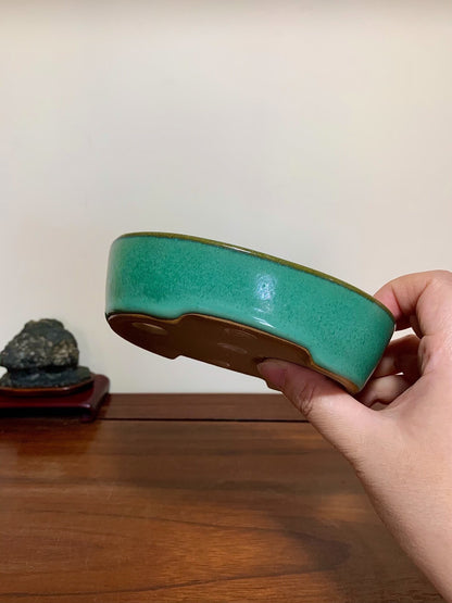 A hand holds a glossy teal glazed pot with a brown unglazed base.