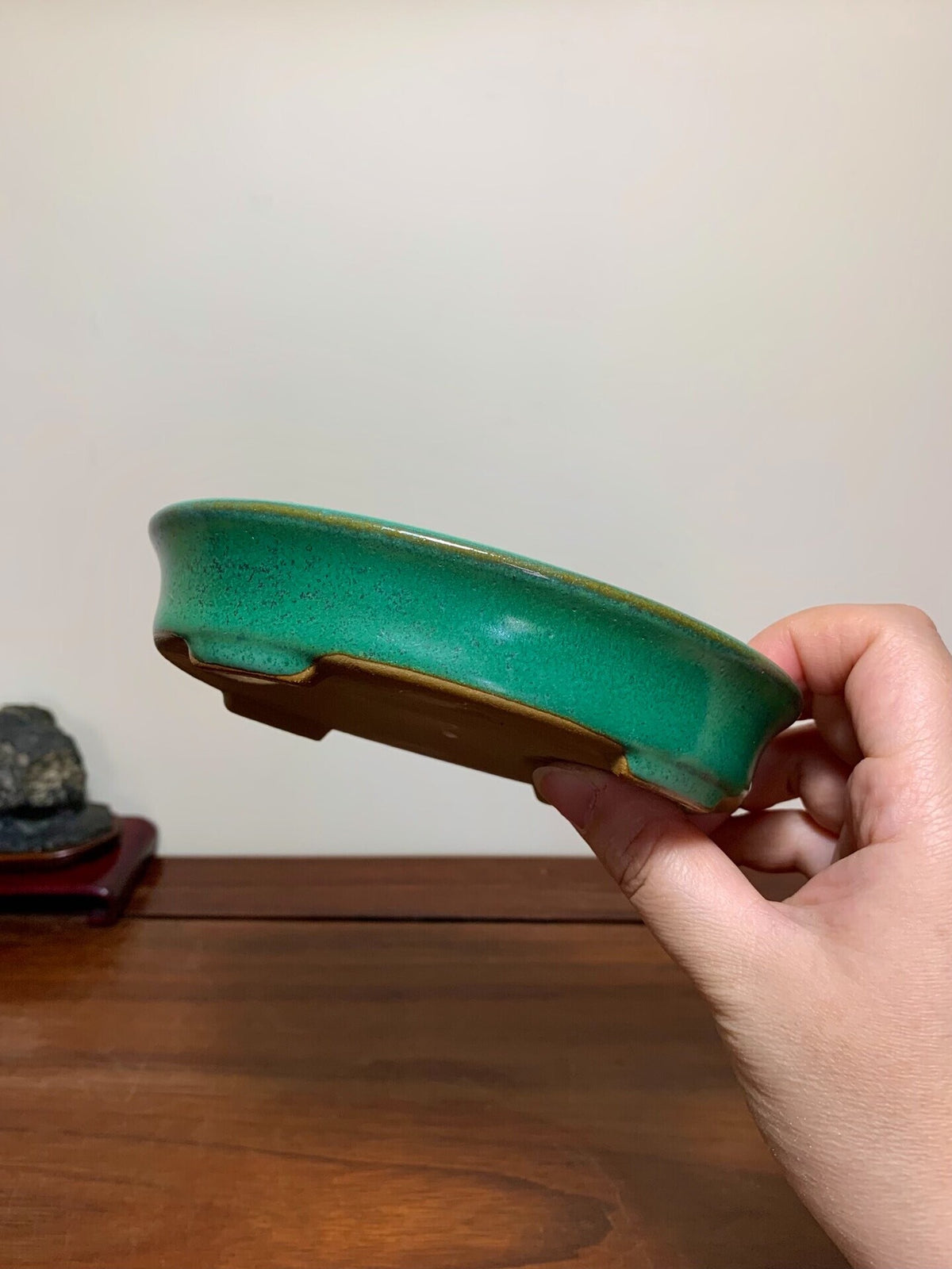 A hand holds a glossy, teal-glazed ceramic bonsai pot with a brown rim and unglazed base.