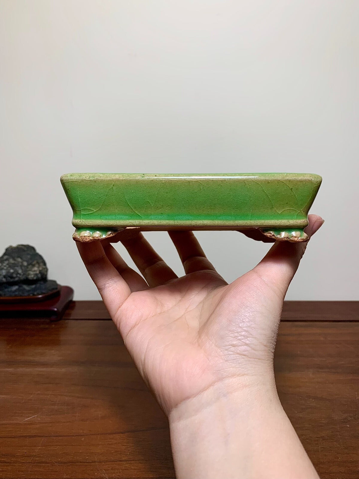 A hand holds a small, rectangular green glazed pot with a crackled surface and gold-trimmed feet.