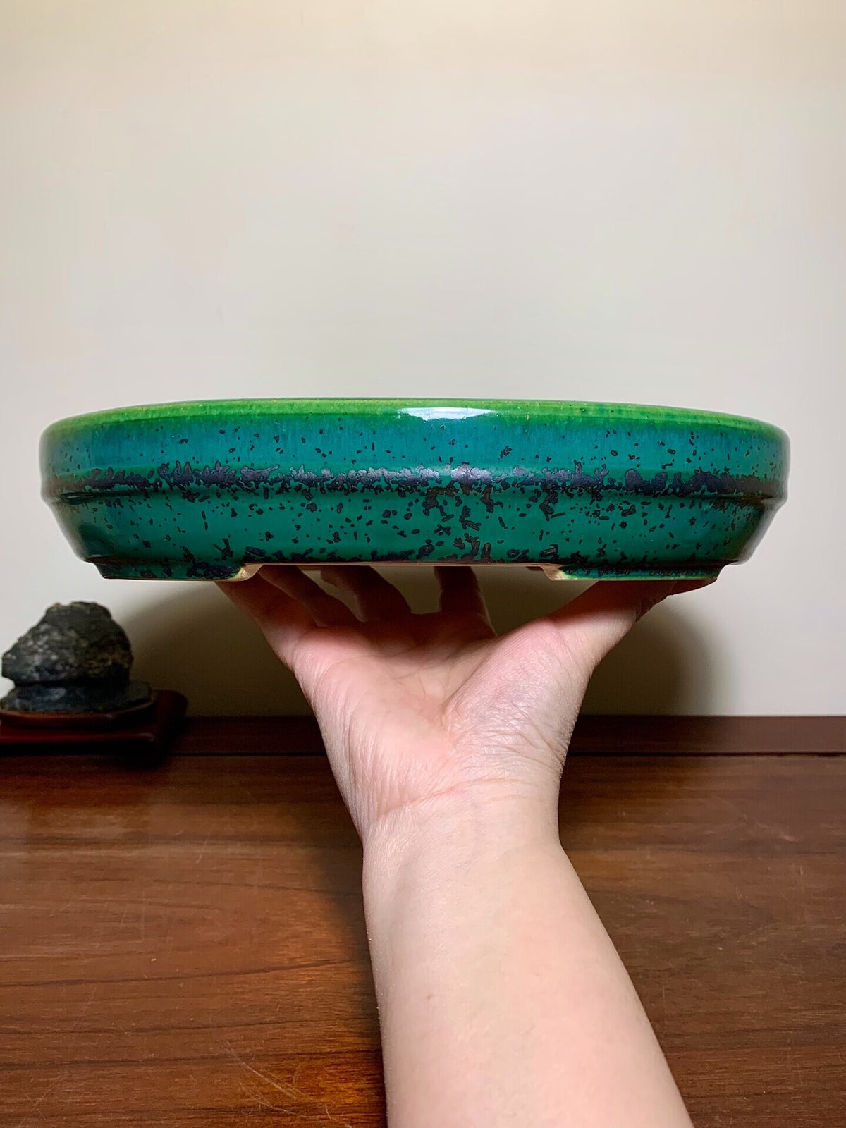 A hand holds a glossy, round, green glazed pot with dark speckles and a wide rim.
