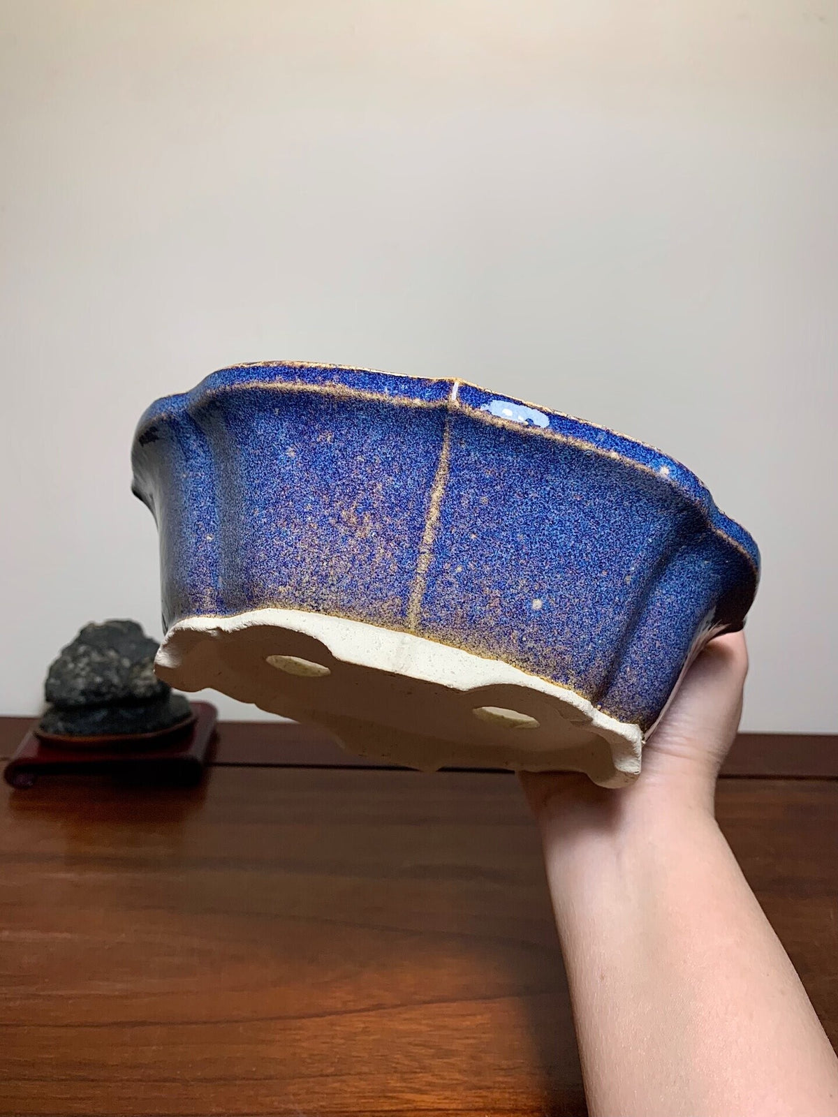 A hand holds a blue speckled glazed pot with a cream-colored base and three drainage holes.
