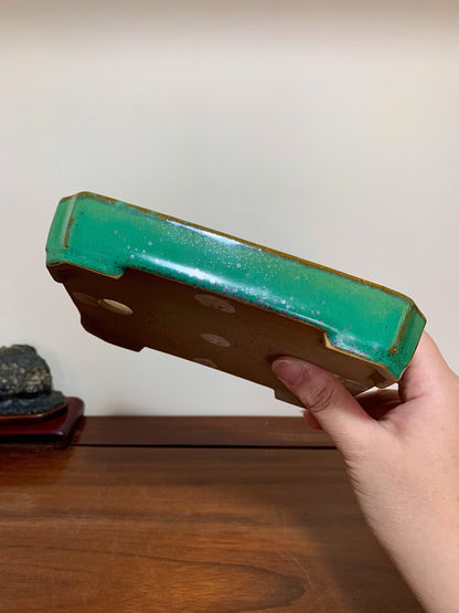A hand holds a rectangular glazed pot with a vibrant green rim and brown body.