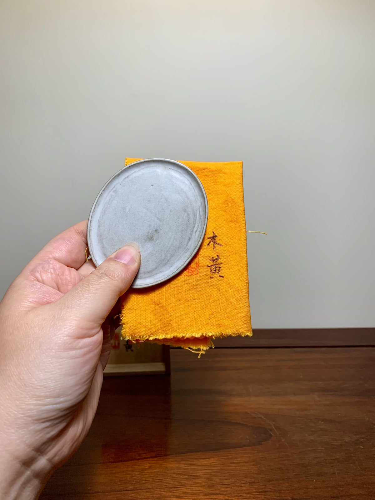 A hand holds a small, round, grey, unglazed ceramic lid over an orange cloth with black Japanese characters.