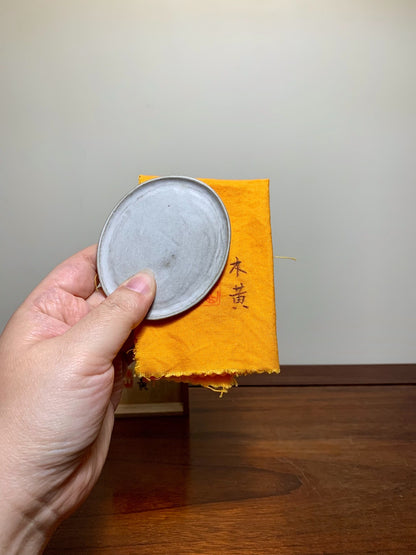 A hand holds a small, round, grey, unglazed ceramic lid over an orange cloth with black Japanese characters.