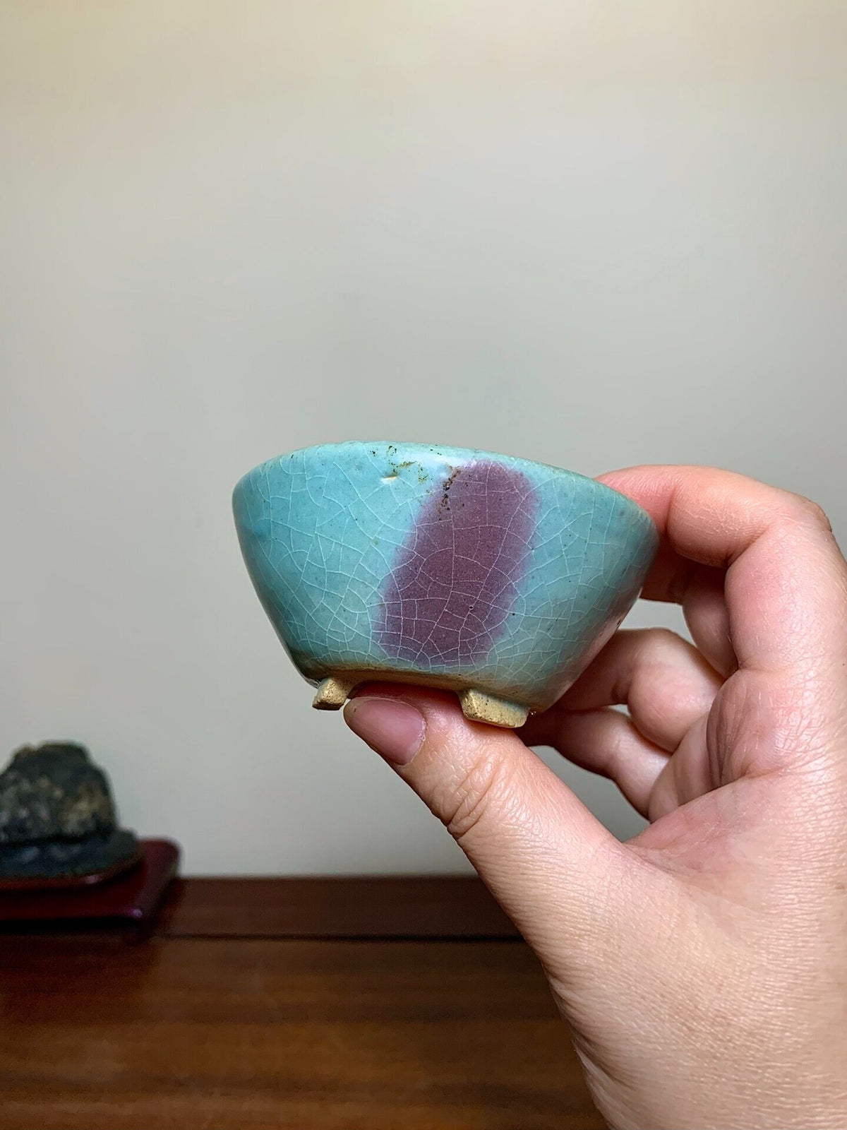 A hand holds a small, crackled-glazed pot with turquoise and mauve coloration and three feet.