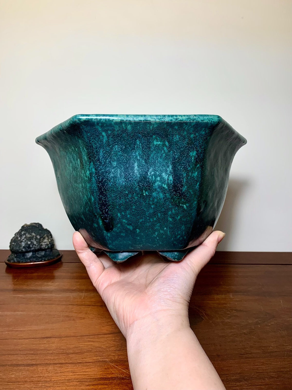 A hand holds a square, teal-glazed pot with a speckled, glossy finish and flared rim.