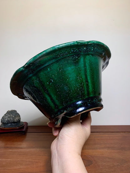A hand holds a glossy, deep green ceramic pot with flared rim and vertical ridges.