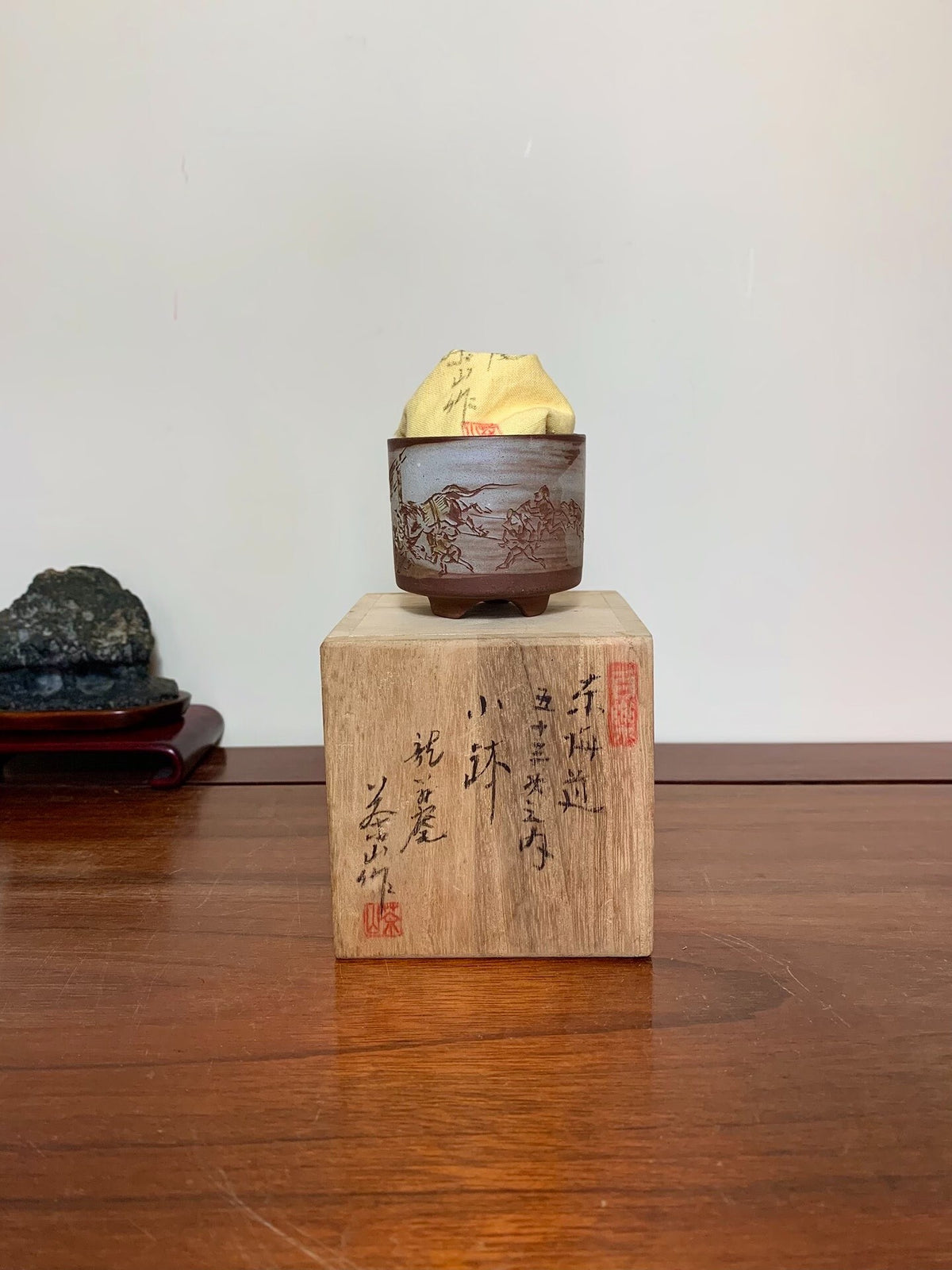 A small brown glazed pot with landscape painting holds a wrapped bonsai, resting on a wooden block with calligraphy.
