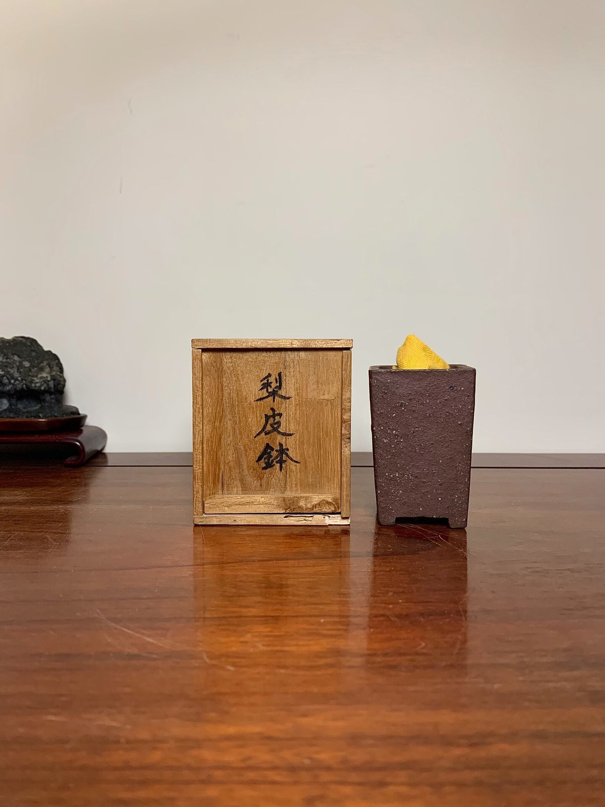 A dark brown, textured ceramic pot holds a yellow object beside a wooden box with black characters.