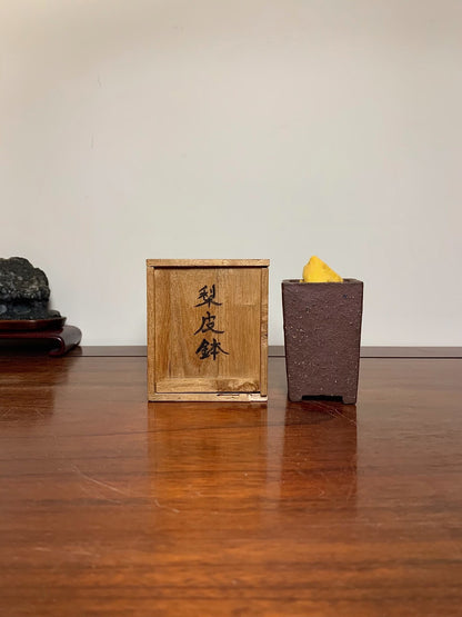 A dark brown, textured ceramic pot holds a yellow object beside a wooden box with black characters.