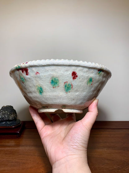 A hand holds a light grey glazed pot with red and green speckles and a scalloped rim.