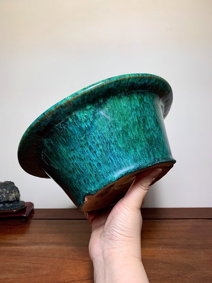 A hand holds a glossy, teal-glazed ceramic pot with a flared rim and brown base.