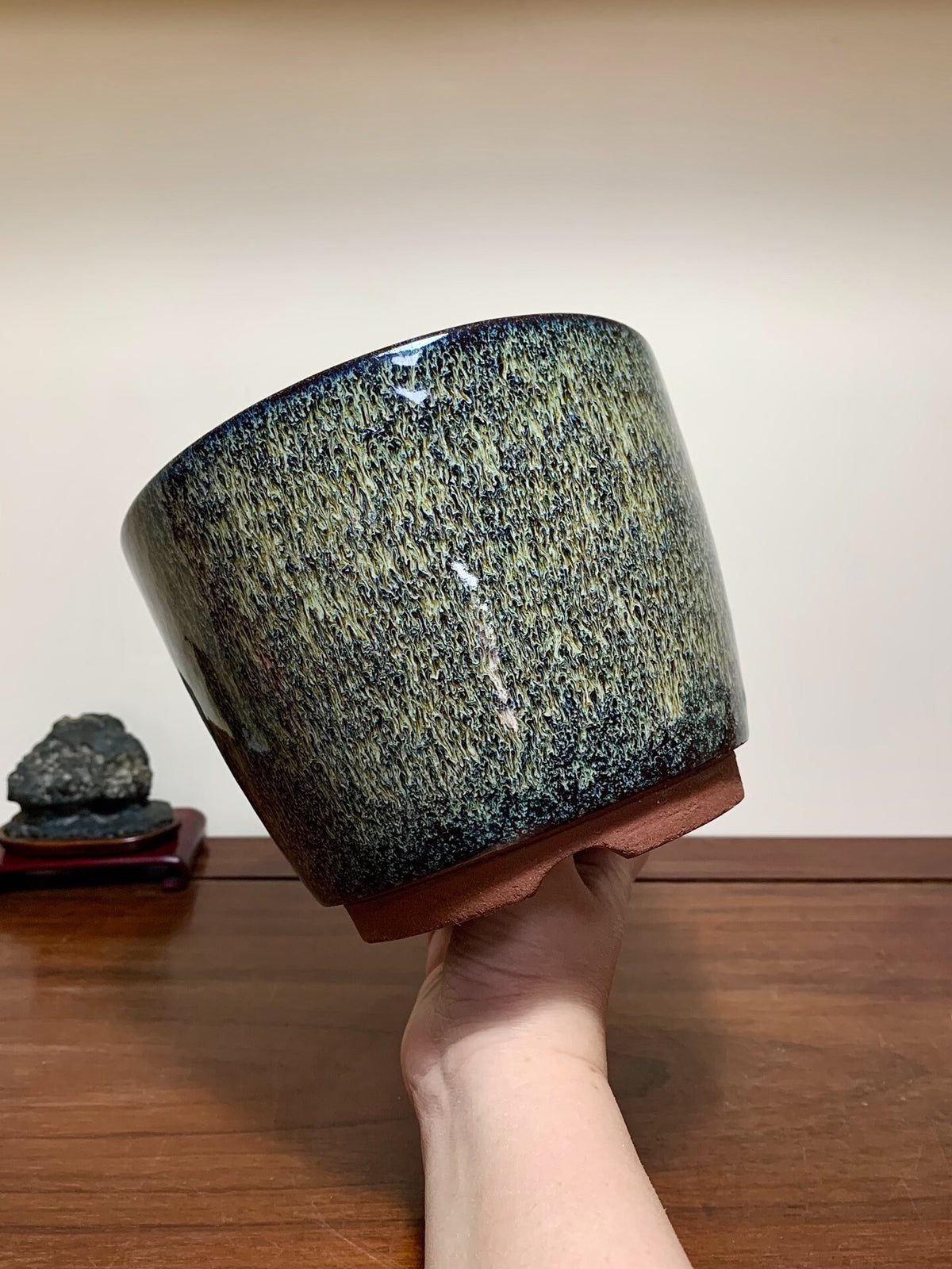 A hand holds a glossy, speckled green and black glazed pot with a terracotta base.