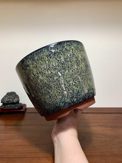 A hand holds a glossy, speckled green and black glazed pot with a terracotta base.