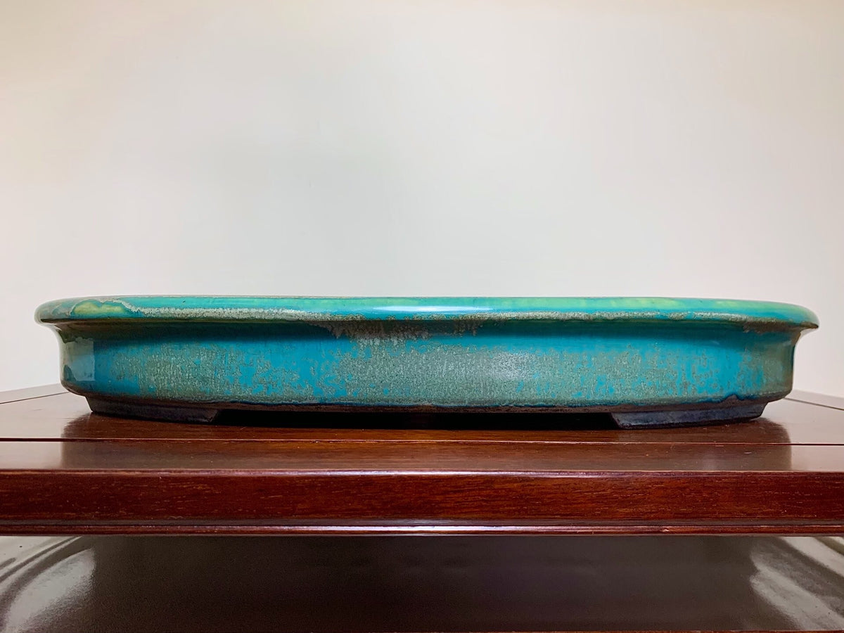 A long, shallow turquoise glazed pot with a worn, speckled surface rests on a wooden shelf.