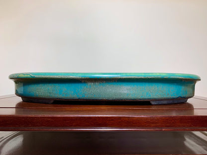 A long, shallow turquoise glazed pot with a worn, speckled surface rests on a wooden shelf.