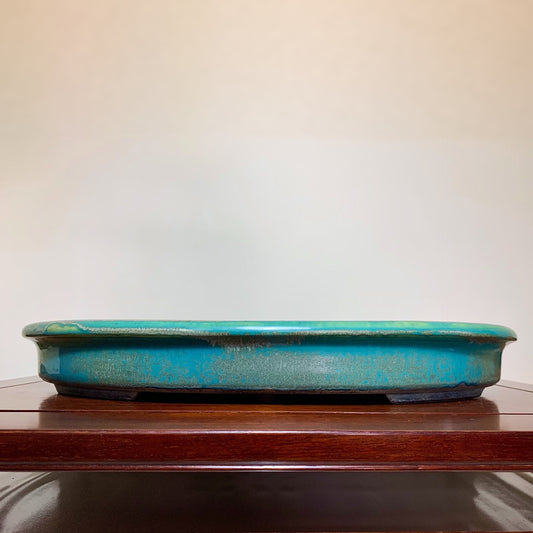 A long, shallow turquoise glazed pot with a worn, speckled surface rests on a wooden shelf.