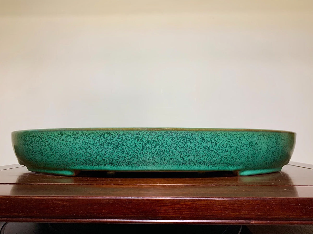 A low, wide, teal-glazed ceramic pot with speckled surface rests on a wooden surface.