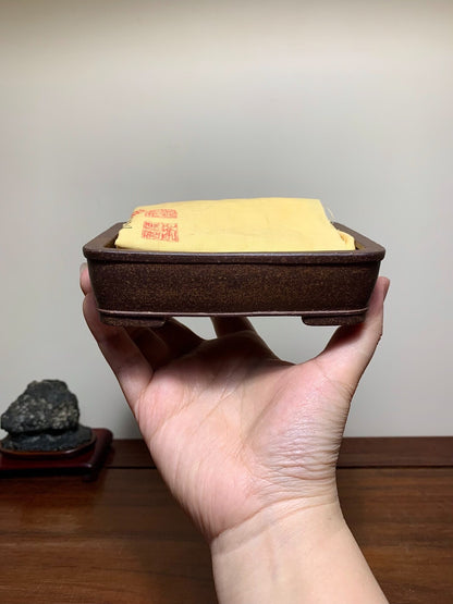 A hand holds a small, dark brown, unglazed rectangular pot with a yellow cloth inside.