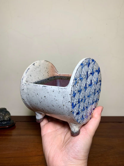 A hand holds a speckled grey ceramic pot with blue fan-patterned sides and three short feet.