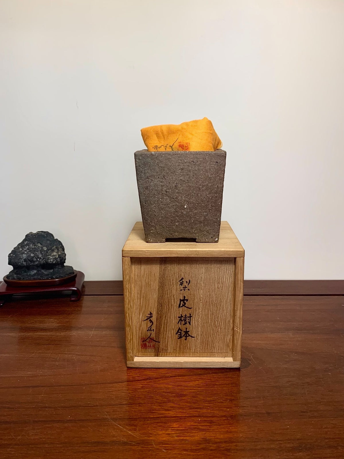 Dark grey unglazed square pot with orange cloth inside, resting on a wooden box with black characters.