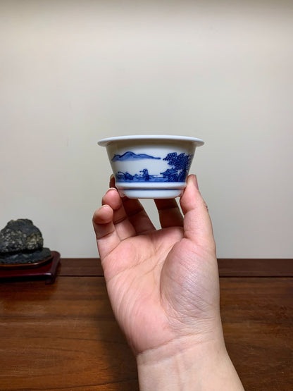 A hand holds a small white ceramic pot with blue landscape painting.