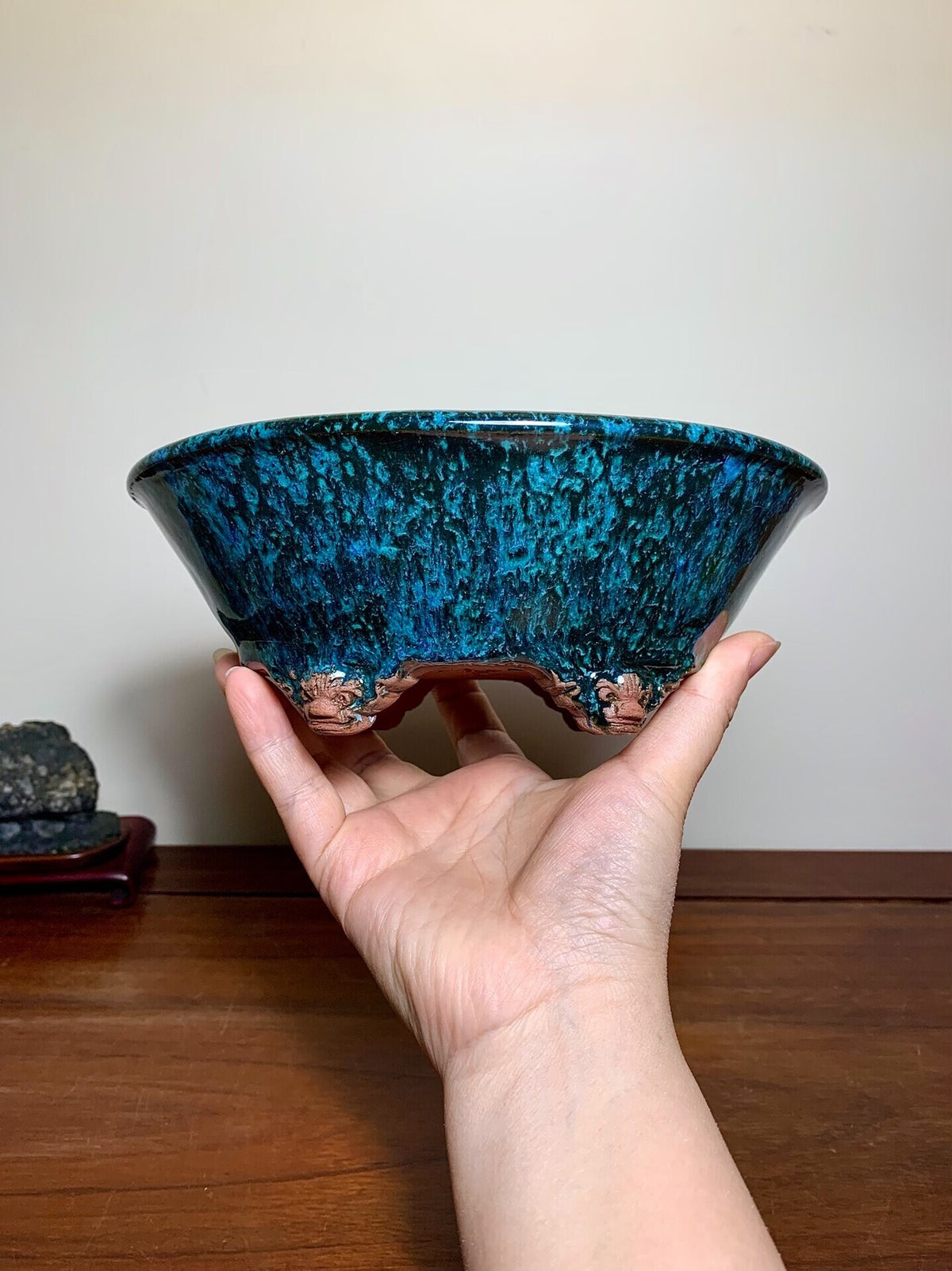 A hand holds a wide, shallow bowl with a glossy, speckled blue glaze and three carved feet.