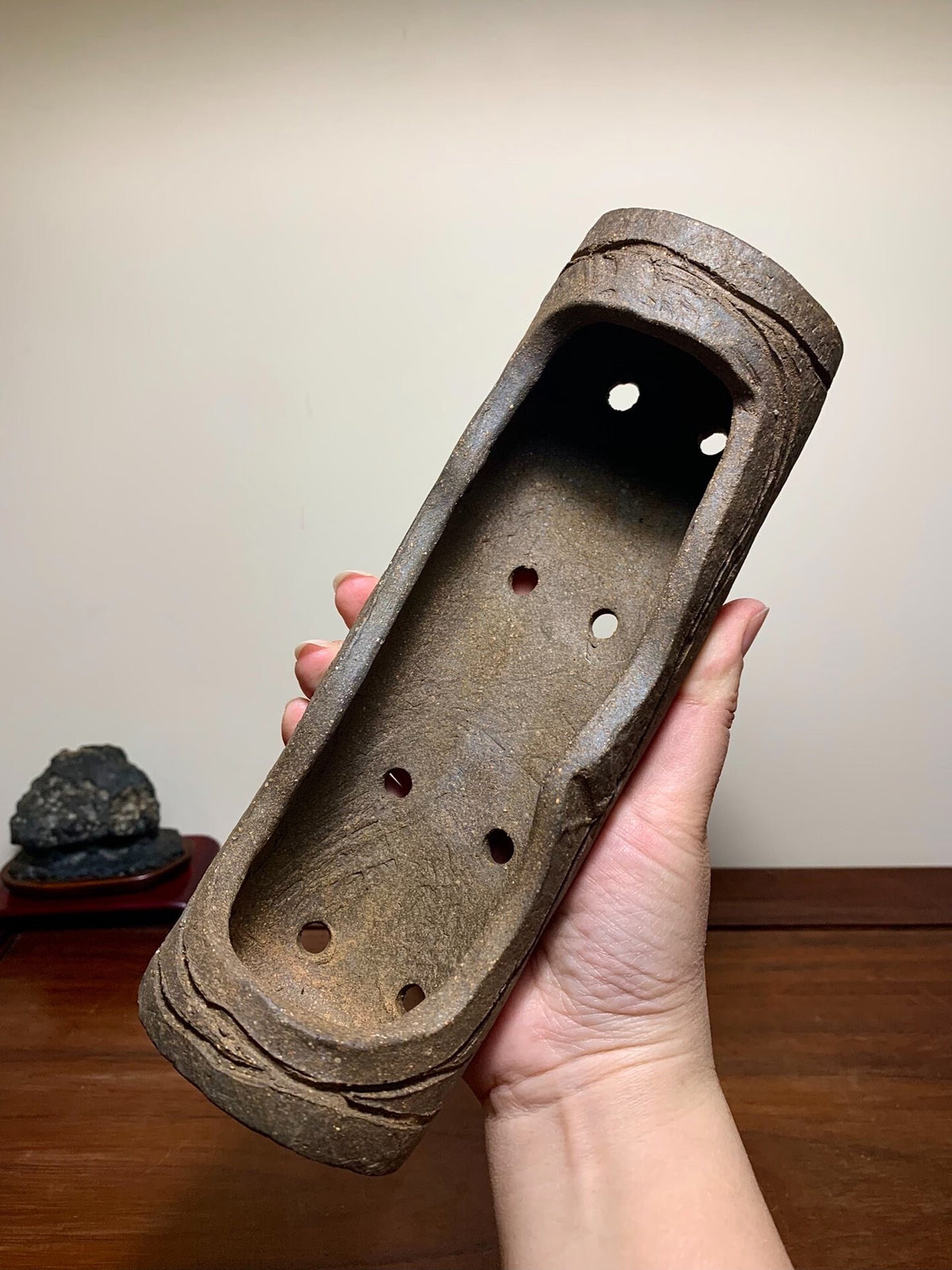 A hand holds a weathered, brown, unglazed rectangular pot with seven holes and rough texture.