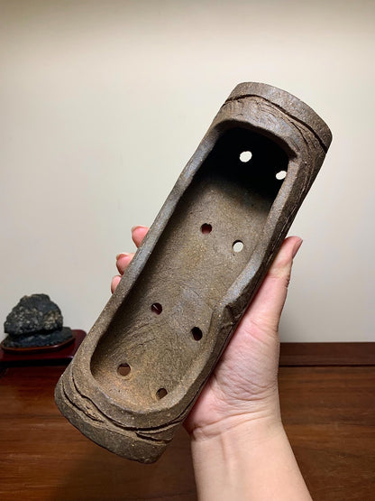 A hand holds a weathered, brown, unglazed rectangular pot with seven holes and rough texture.