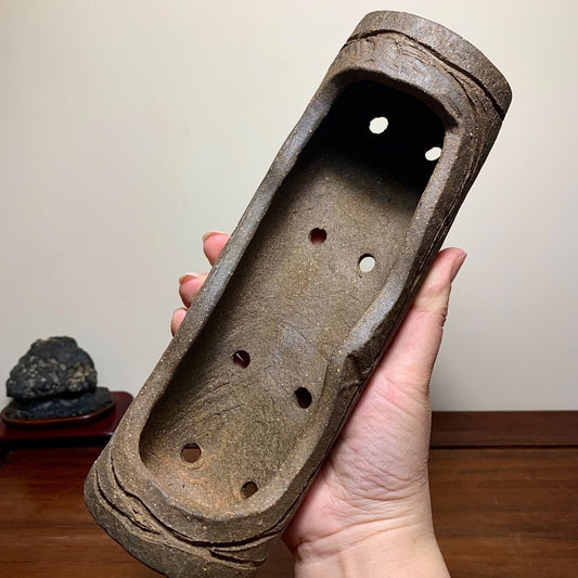 A hand holds a weathered, brown, unglazed rectangular pot with seven holes and rough texture.