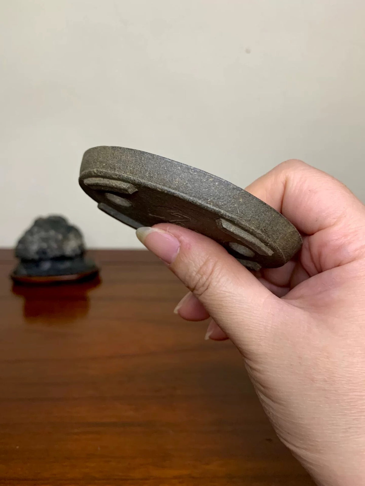 A hand holds a dark grey, unglazed, circular bonsai pot base with three raised feet.