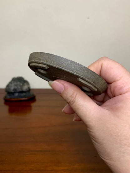 A hand holds a dark grey, unglazed, circular bonsai pot base with three raised feet.