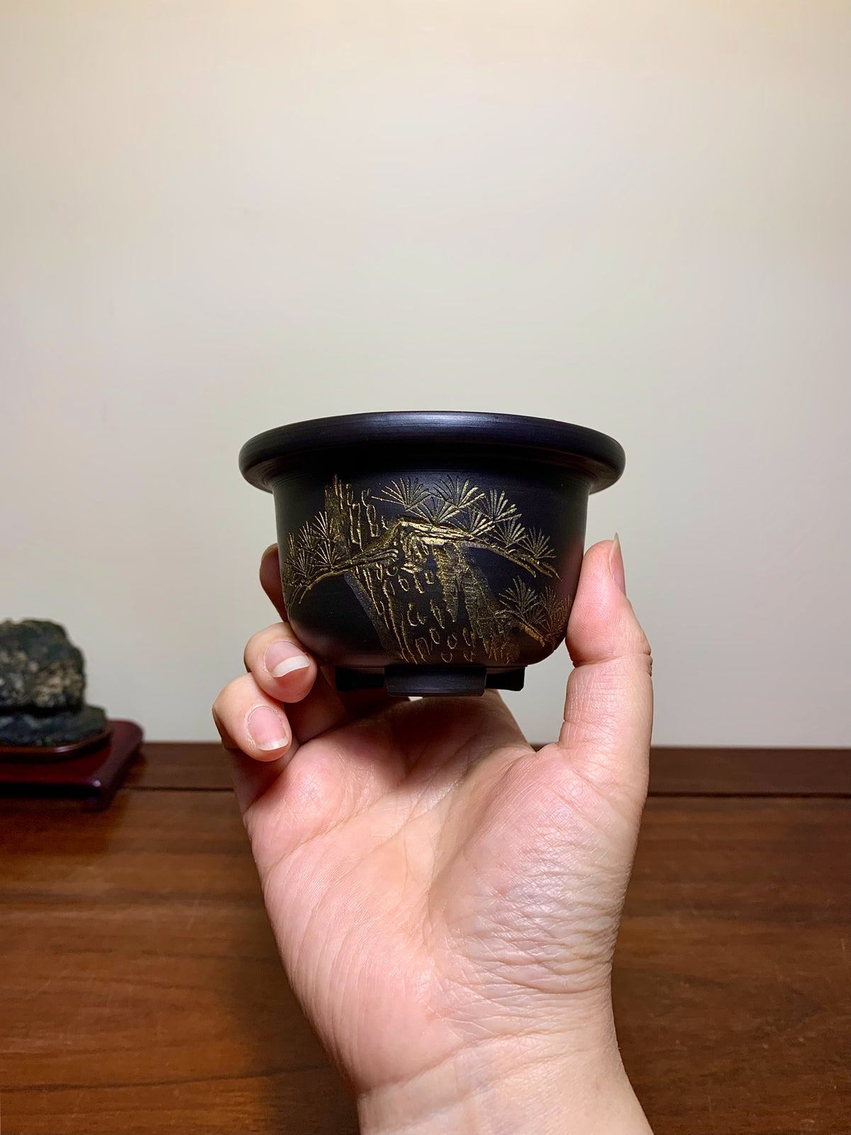 A hand holds a small, dark glazed pot with gold-leaf pine tree decoration.