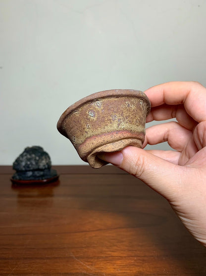 A hand holds a small, brown, unglazed ceramic pot with speckled surface and uneven rim.