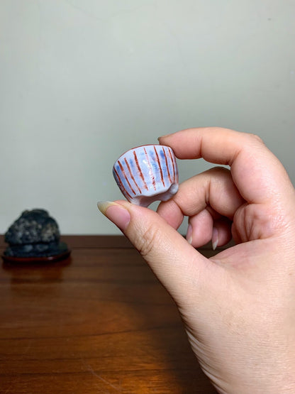 A hand holds a small, white glazed pot with red diagonal stripes.