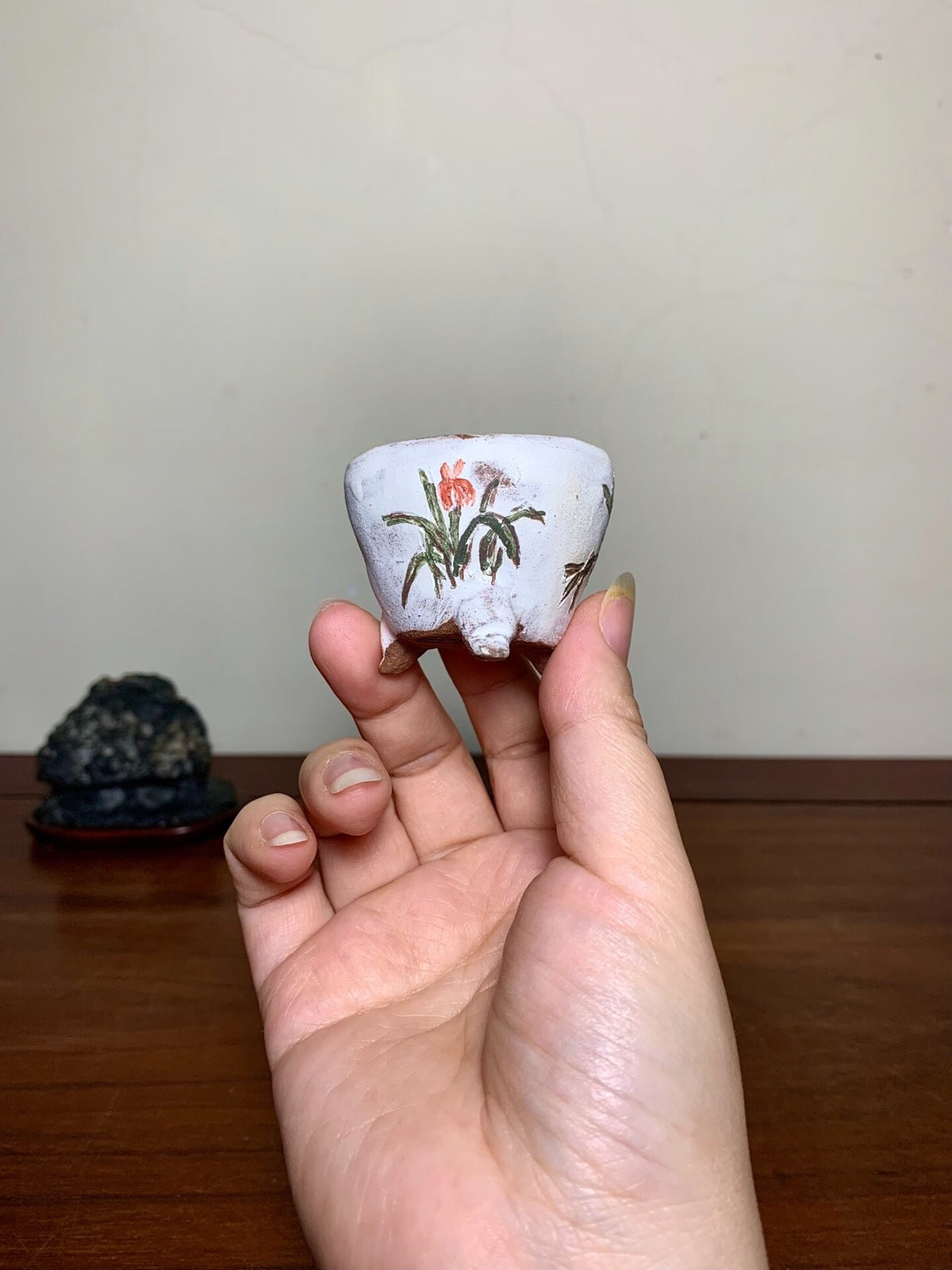 A hand holds a small, white, glazed pot with a painted red flower and green leaves.