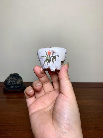 A hand holds a small, white, glazed pot with a painted red flower and green leaves.